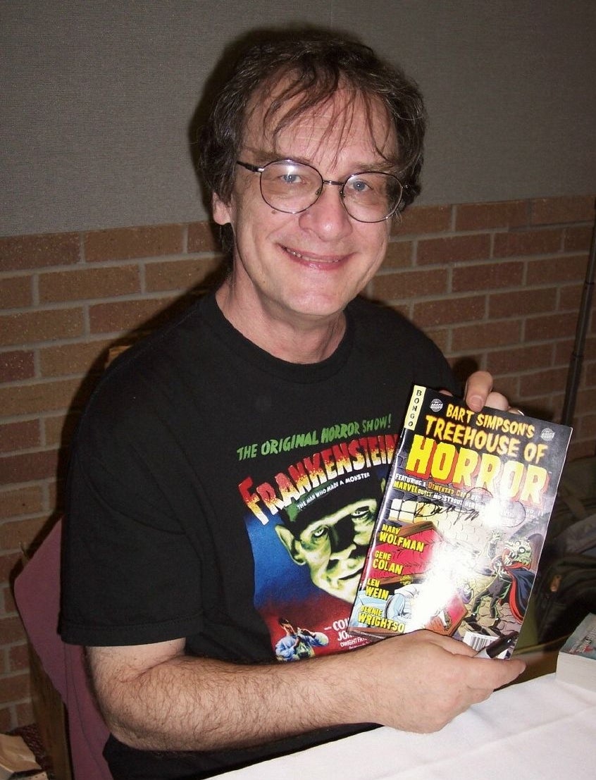 A smiling man with glasses wearing a Frankenstein t-shirt holds up a copy of the Bart Simpson's Treehouse of Horror comic book at a table.